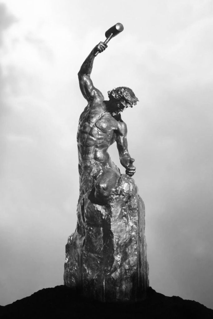 A dramatic black and white sculpture of a muscular figure holding a hammer, poised on a rocky pedestal, symbolizing strength and creation.